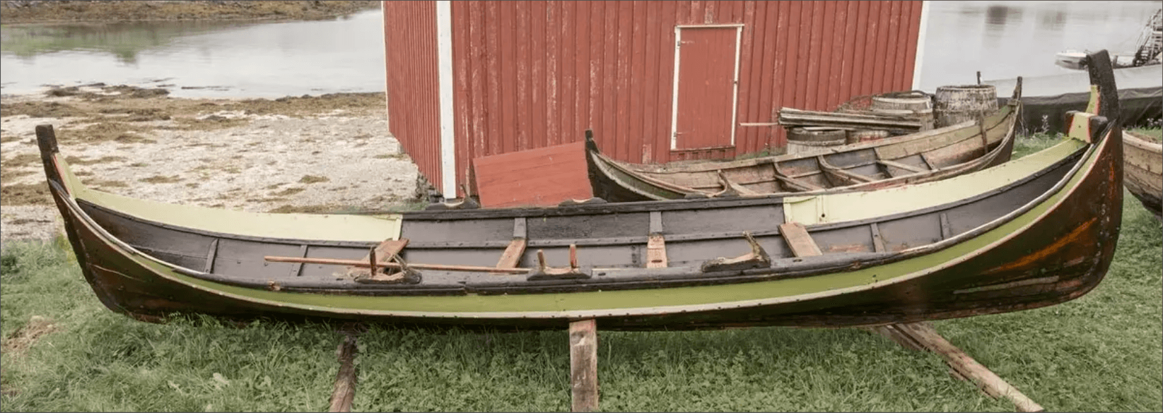 Long traditional wooden boat with green gunwales displayed beside a red boathouse on Herøy