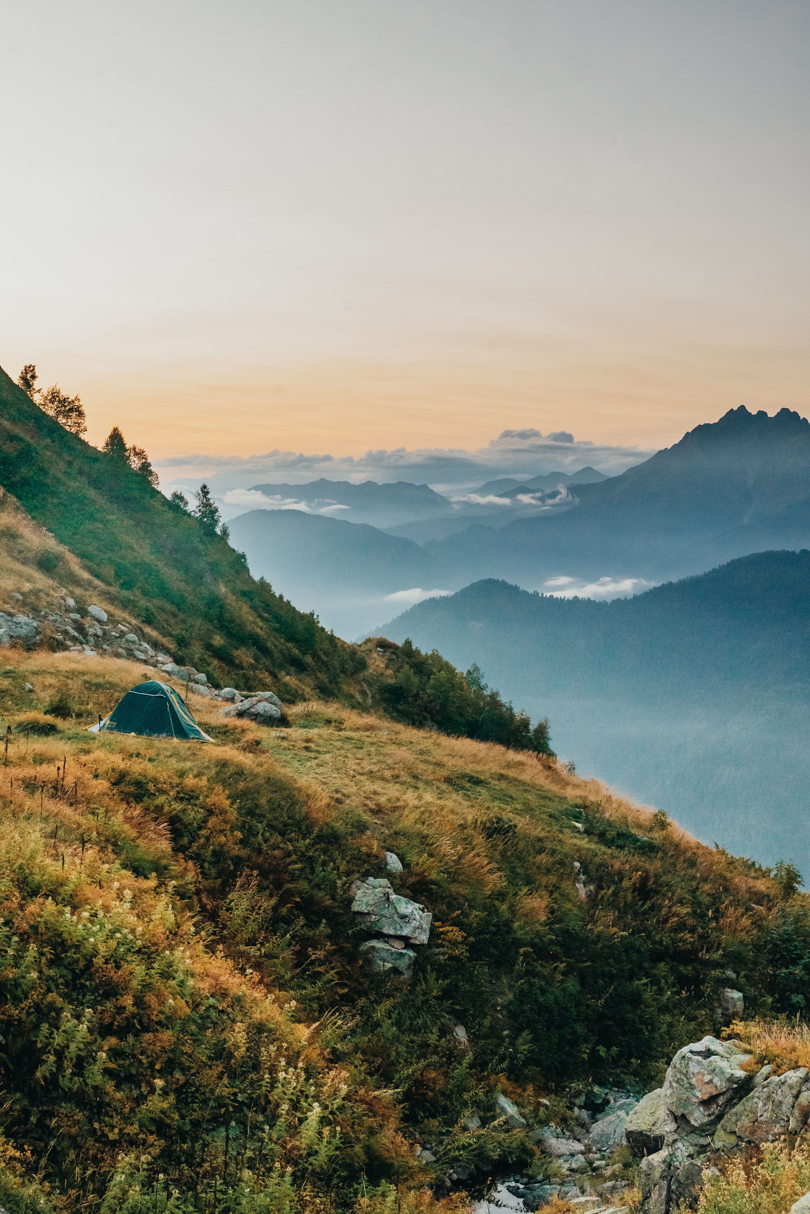 A small tent on a grassy mountain hillside above layers of Norwegian peaks at dusk